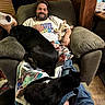 armchair, beard, black_cat, black_dog, blanket, casual_clothing, cat, cozy, dog, home, indoors, man, pet, relaxing, sleeping_cat, sleeping_dog, small_dog, smiling, white_dog, wood_floor