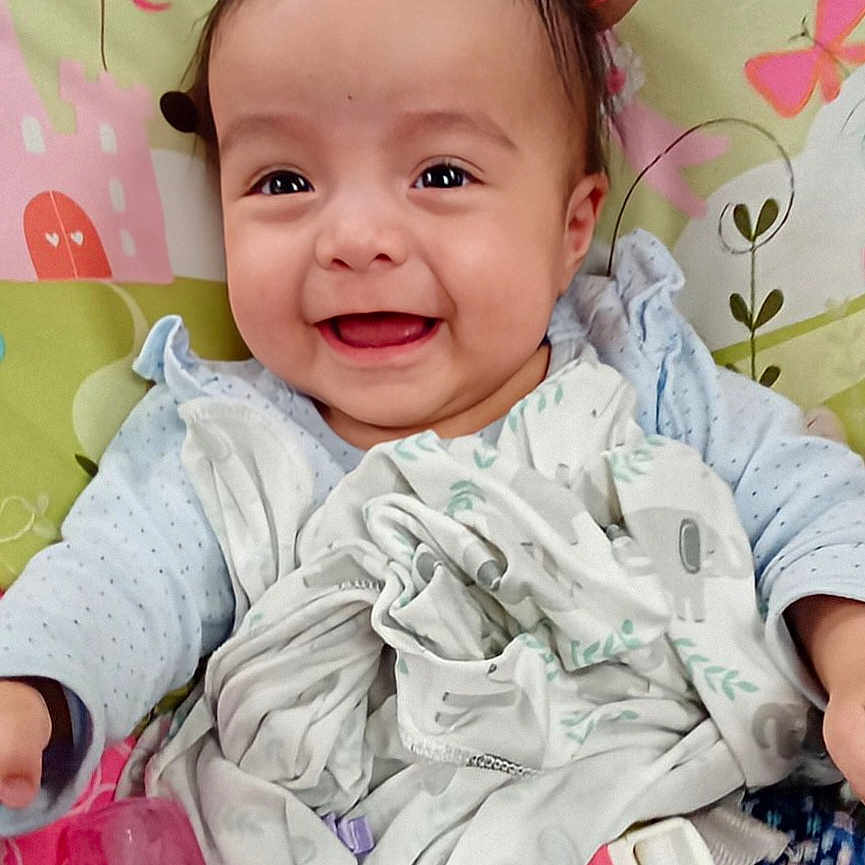 Delilah is registered to the contest to win money with this photo: baby, bear_ears, blanket, bottle, child, clothing, colorful, cute, face, happy, head, indoors, infant, lying_down, patterned_background, person, playful, smiling, soft_toys, young_child