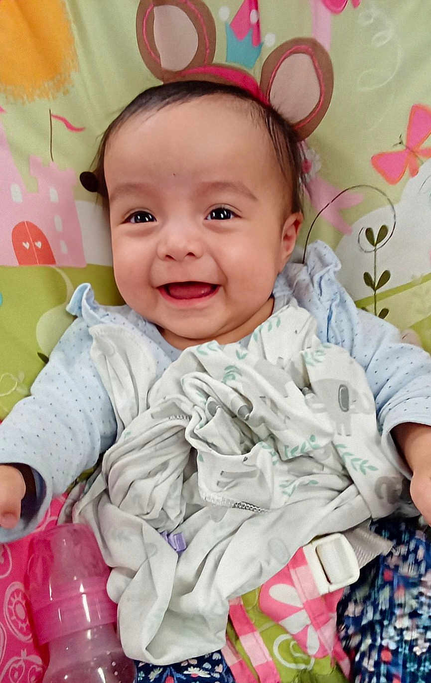 Delilah is registered to the contest to win money with this photo: baby, smiling, face, bear_ears, blanket, bottle, child, infant, cute, happy, patterned_background, clothing, lying_down, indoors, colorful, soft_toys, head, person, young_child, playful