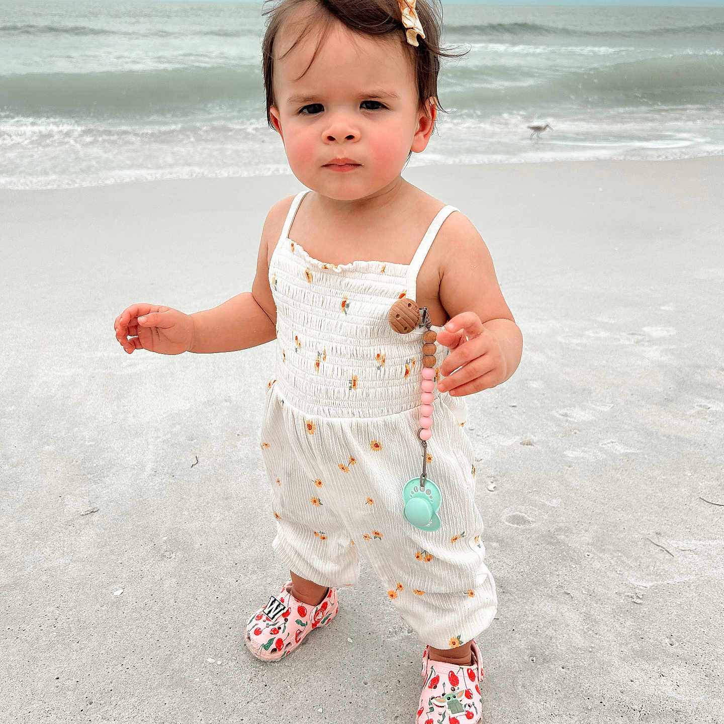 Winter is registered to the contest to win money with this photo: beach, child, cloudy_sky, cute, expression, footwear, hair_clip, nature, ocean, outdoor, pacifier, person, romper, sand, standing, summer, toddler, vacation, water, waves