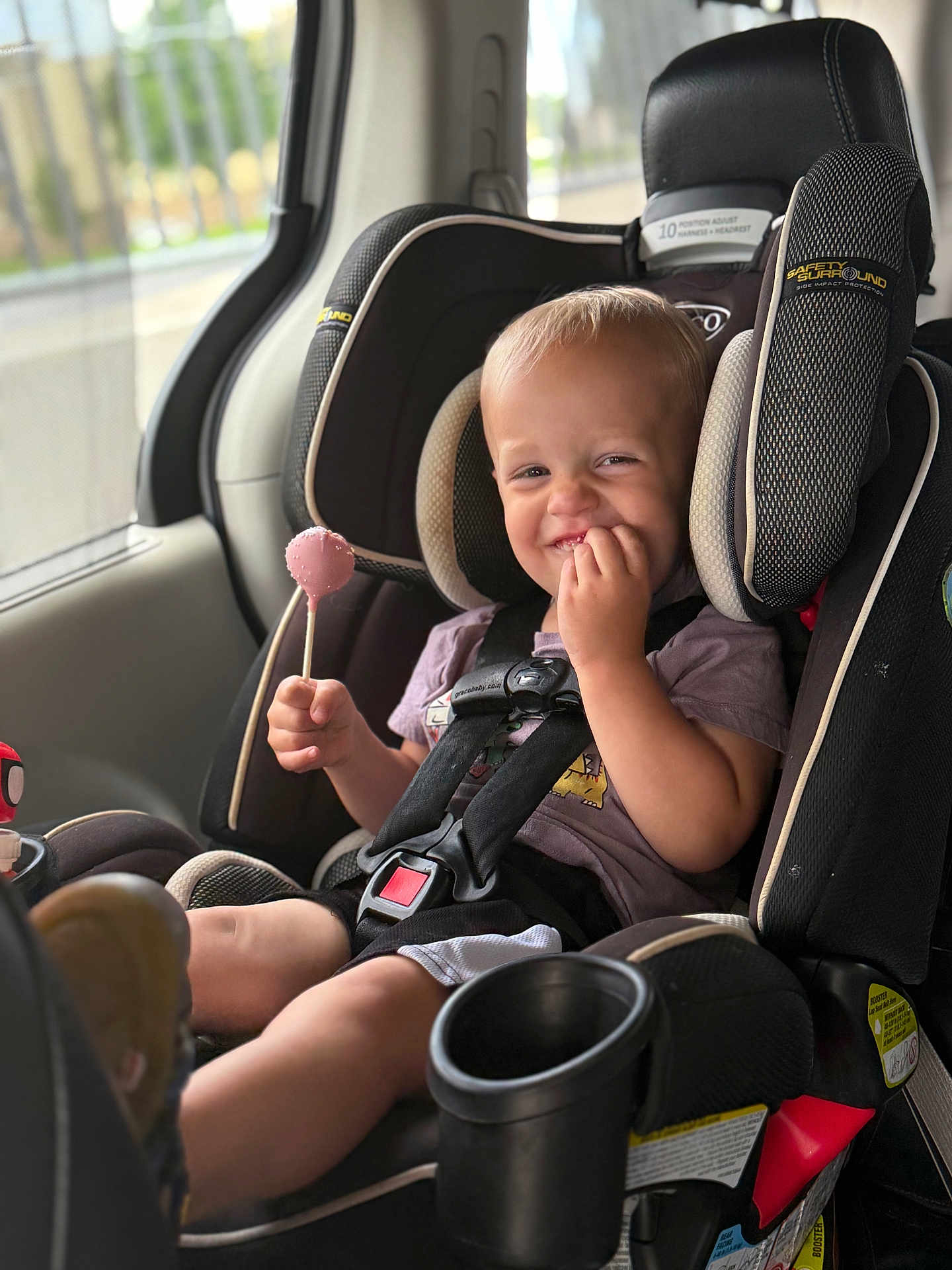 Graynger is registered to the contest to win money with this photo: toddler, child, car_seat, lollipop, smile, happy, seatbelt, vehicle_interior, window, daylight, casual_clothing, safety_harness, black_car_seat, cute, portrait, young_child, seat_cup_holder, car, sitting, fun