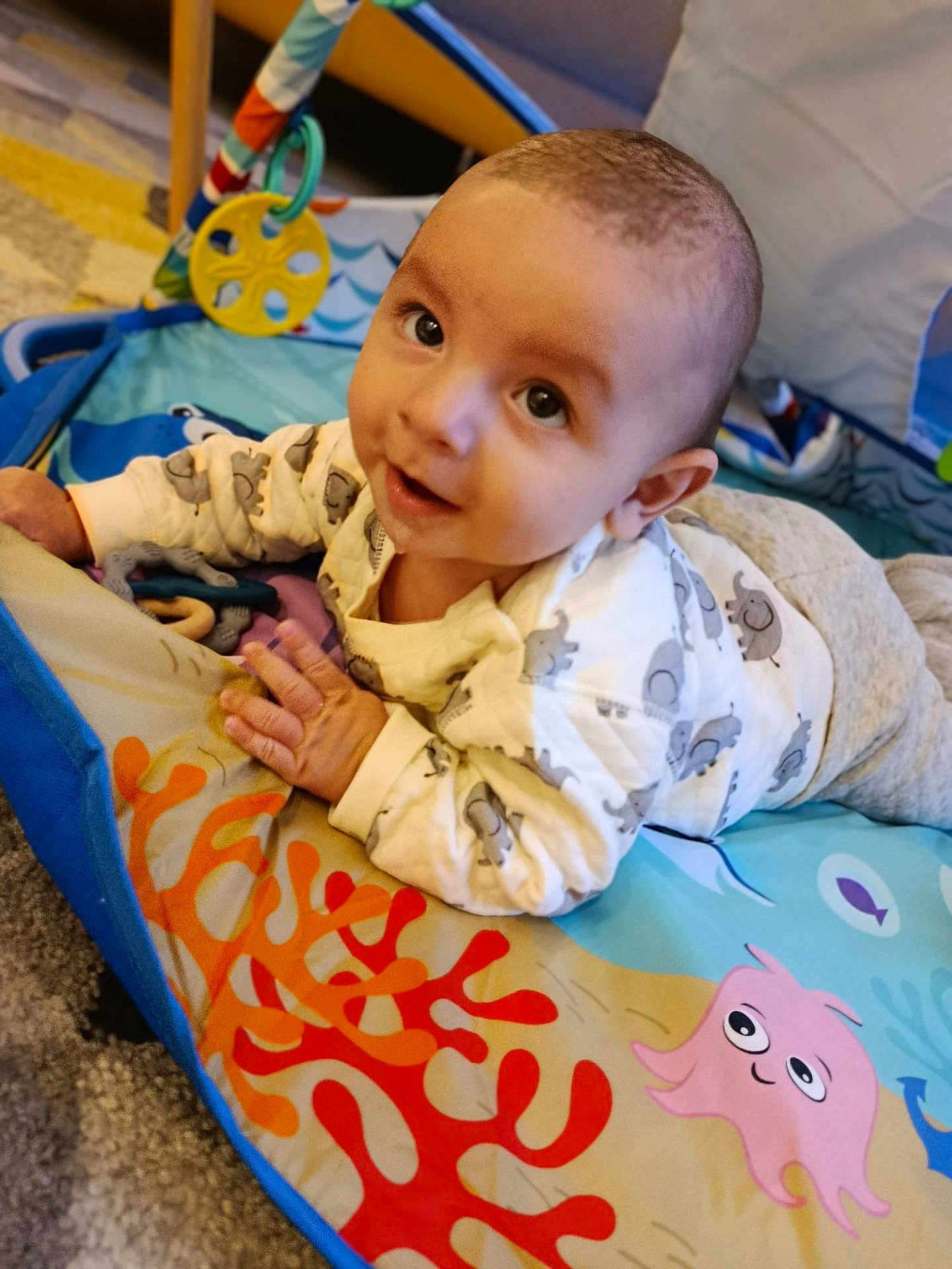 Sully a rejoint le concours — aidez-le/la à gagner de superbes lots ! baby, child, infant, play_mat, elephant_print, smile, face, clothing, toy, floor, carpet, indoor, cute, person, toddler, happy, colorful, pattern, lying_down, curious