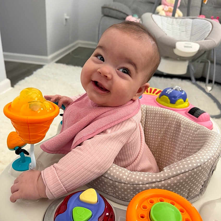 Madelyn joined the competition — help win amazing prizes! baby, smiling, pink_clothing, activity_center, toy, indoor, living_room, baby_swing, carpet, furniture, child, cute, happy, face, infant, play, seat, home, person, portrait