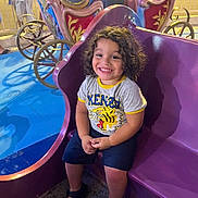 Ethan participe au concours pour gagner de l'argent avec cette photo : child, smiling, curly_hair, purple_carriage, amusement_park, ride, people, night, colorful, happy, casual_clothing, sandals, crowd, outdoor, fun, entertainment, portrait, seated, festival, family