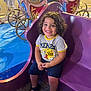 child, smiling, curly_hair, purple_carriage, amusement_park, ride, people, night, colorful, happy, casual_clothing, sandals, crowd, outdoor, fun, entertainment, portrait, seated, festival, family