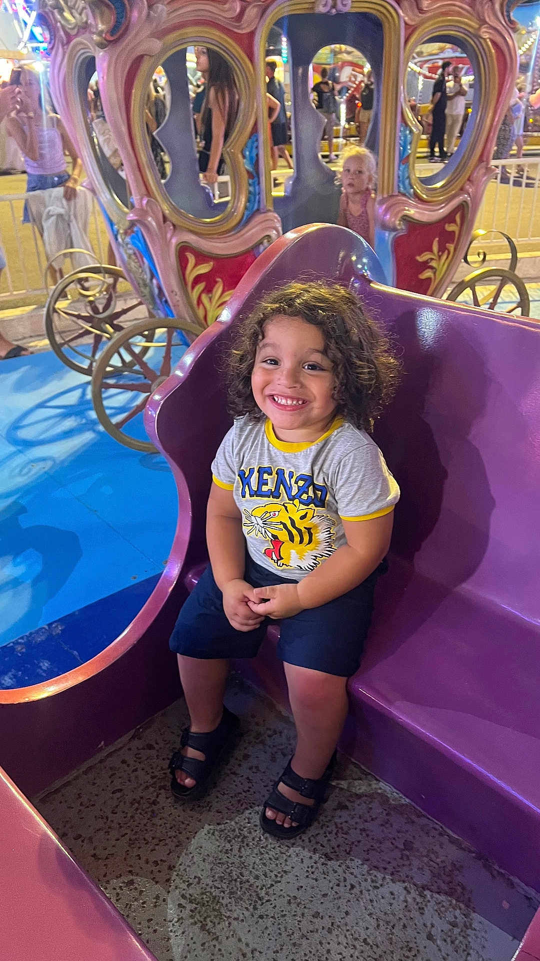 Ethan participe au concours pour gagner de l'argent avec cette photo : child, smiling, curly_hair, purple_carriage, amusement_park, ride, people, night, colorful, happy, casual_clothing, sandals, crowd, outdoor, fun, entertainment, portrait, seated, festival, family
