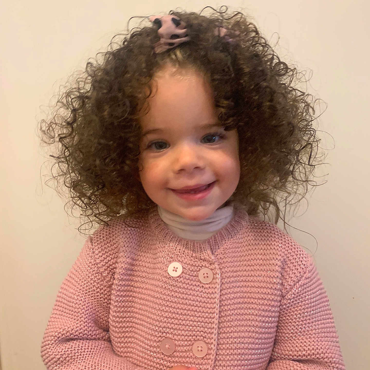 Késia participe au concours pour gagner de l'argent avec cette photo : buttoned_sweater, child, curly_hair, cute, cute_expression, face, fashion, hands_clasped, happy, indoor, innocent, light_background, long_sleeves, person, pink_sweater, portrait, smiling, toddler, warm_clothing, young
