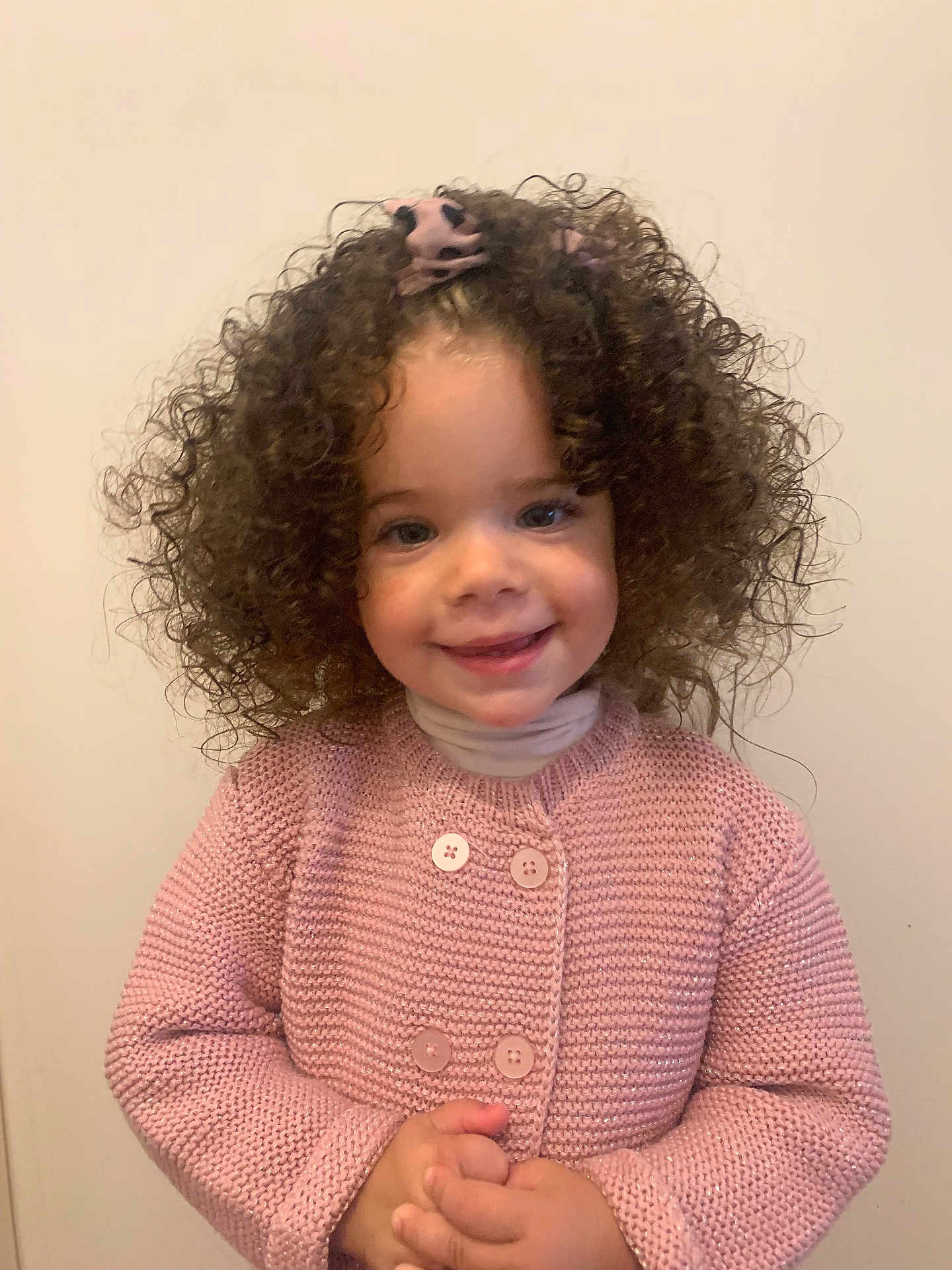 Késia participe au concours pour gagner de l'argent avec cette photo : child, toddler, curly_hair, pink_sweater, smiling, portrait, indoor, hands_clasped, cute, young, person, face, happy, buttoned_sweater, long_sleeves, light_background, fashion, cute_expression, warm_clothing, innocent