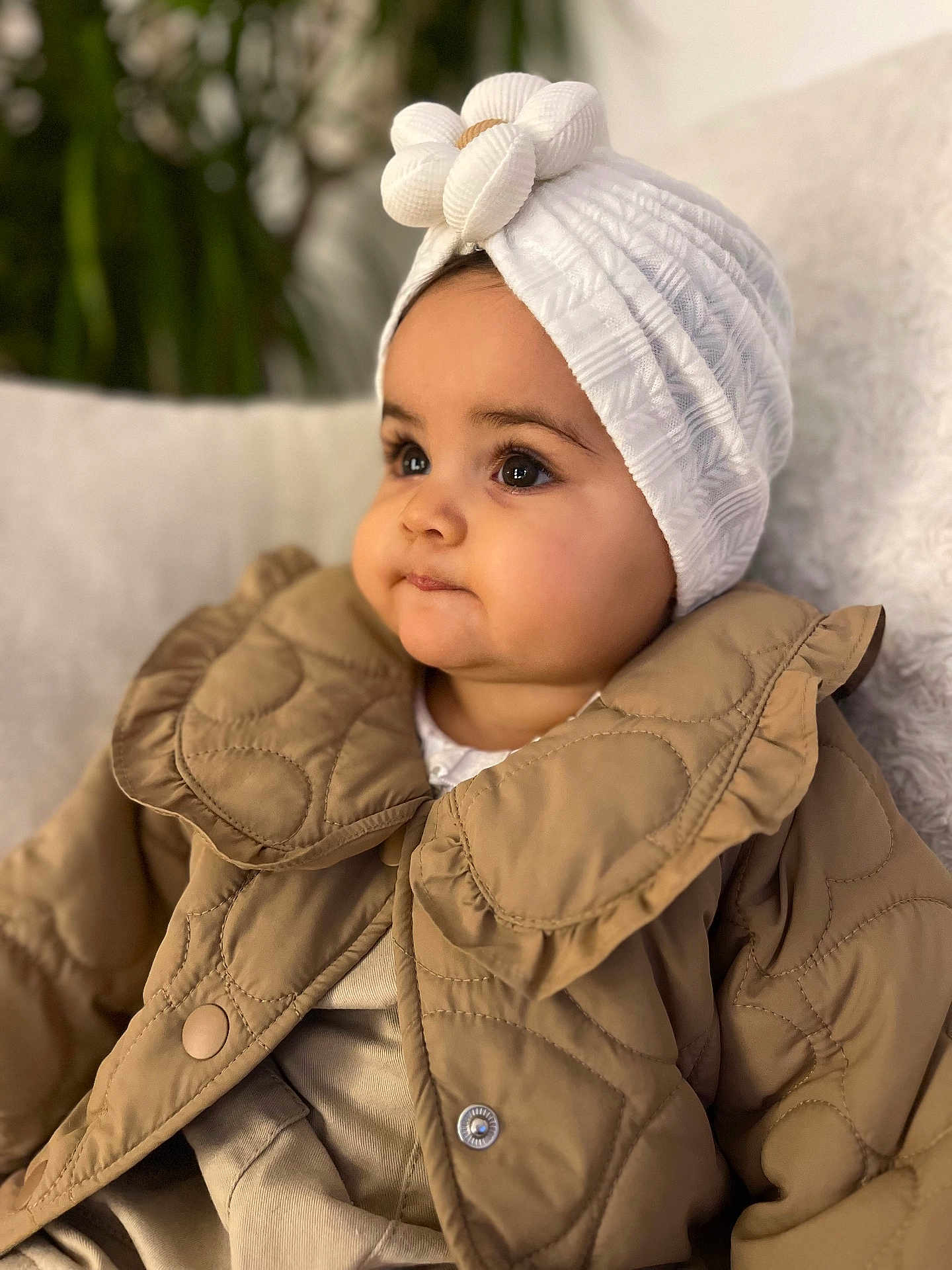 Agathe participe au concours pour gagner de l'argent avec cette photo : baby, infant, headwrap, white_hat, quilted_jacket, brown_coat, chubby_cheeks, big_eyes, portrait, close_up, indoor, couch, blanket, plant, sitting, fashion, cute, eyelashes, soft_lighting, warm_colors