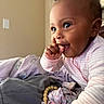 Hilani is registered to the contest to win money with this photo: baby, bedroom, blanket, child, closeup, cozy, curly_hair, cute, face, happy, indoors, infant, onesie, person, pink_clothing, playful, smiling, soft_lighting, thumb_in_mouth, toy