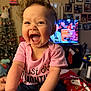 baby, child, smiling, happy, curly_hair, pink_shirt, indoor, christmas_tree, holiday_decor, television, family_room, portrait, candid, warm_lighting, celebration, infant, sitting, joyful, clothing, person