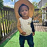 toddler, child, hat, straw_hat, outdoor, deck, wooden_rail, green_carpet, jeans, long_sleeve_shirt, two_tone_clothing, sunny_day, leafy_background, person, footwear, black_shoes, nature, sky, clouds, casual_wear
