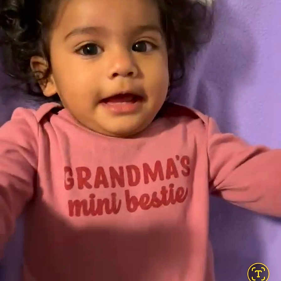 Sitara is registered to the contest to win money with this photo: toddler, child, curly_hair, pink_shirt, text_on_clothing, purple_blanket, lying_down, face, portrait, cute, indoors, baby, smiling, young_child, person, infant, casual_photo, phone_screenshot, clothing, expression