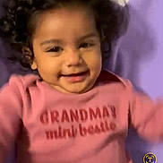 Sitara is registered to the contest to win money with this photo: toddler, child, smiling, curly_hair, pink_shirt, text_on_clothing, purple_blanket, indoor, portrait, happy, baby, cute, person, face, young_child, lying_down, clothing, head, skin, playful