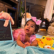 Sitara joined the competition — help win amazing prizes! baby, child, pink_dress, bow, blanket, plush_toy, yellow_tiger, pink_bunny, chalkboard, indoor, floor, curious, smiling, cute, person, toddler, furniture, plant, cozy, portrait