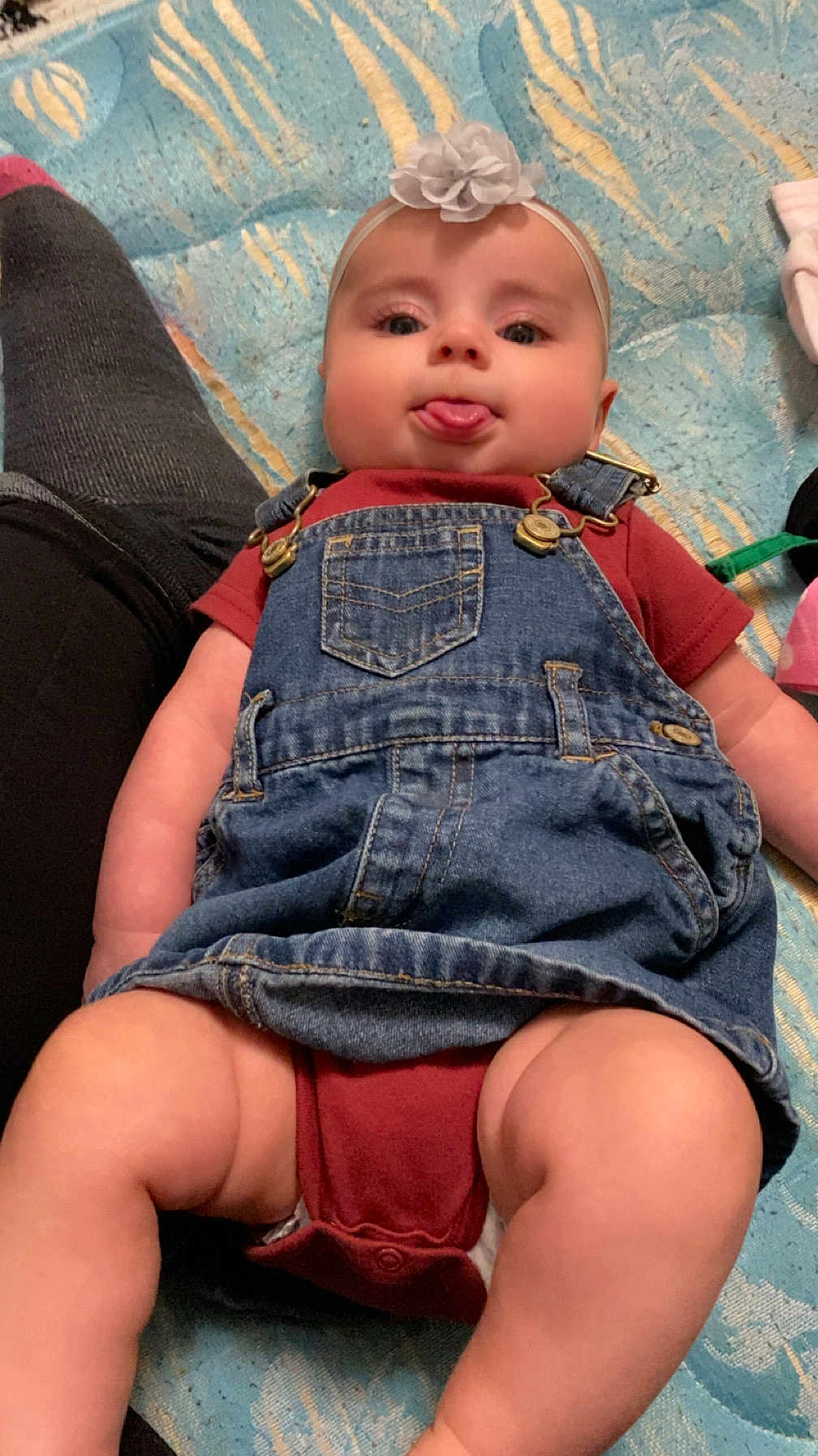 Kylynn joined the competition — help win amazing prizes! baby, baby_clothes, blanket, casual_clothing, child, closeup, cute, denim_overalls, headband, human, indoor, infant, legs, lying_down, pattern, person, playful, skin, smiling, tongue_out