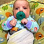 baby, pacifier, blanket, pajamas, cushion, owl_pattern, infant, face, hands, blue_eyes, colorful_clothing, indoor, cozy, soft_texture, child, cute, wrapped, lying_down, closeup, portrait