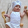 baby, infant, chef_hat, apron, kitchen_utensils, whisk, wooden_spatula, eggs, rolling_pin, smiling, cute, lying_down, white_clothing, patterned_background, cooking_theme, small_hands, playful, portrait, indoors, child