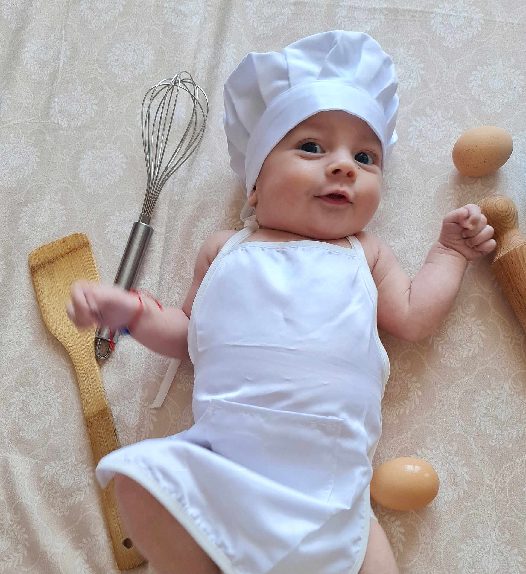 Filip a rejoint le concours — aidez-le/la à gagner de superbes lots ! baby, infant, chef_hat, apron, kitchen_utensils, whisk, wooden_spatula, eggs, rolling_pin, smiling, cute, lying_down, white_clothing, patterned_background, cooking_theme, small_hands, playful, portrait, indoors, child