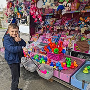 Robert participe au concours pour gagner de l'argent avec cette photo : child, boy, smiling, thumbs_up, carnival_game, plastic_ducks, fishing_game, stuffed_animals, outdoor, fair, toys, colorful, play, casual_clothing, jacket, sneakers, basket, street, fun, happy