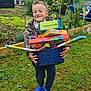 child, boy, smiling, outdoor, garden, grass, toys, basket, blue_crocs, jacket, playful, happy, nature, plants, greenery, casual_clothing, daylight, fun, holding, portrait