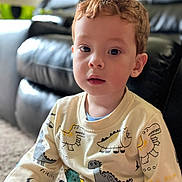 John Paul joined the competition — help win amazing prizes! bright_eyes, casual_clothing, child, couch, curious, curly_hair, cute, dinosaur, expression, face, home, indoor, living_room, person, portrait, seated, soft_lighting, sweater, toddler, young_child