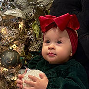 Ainara is registered to the contest to win money with this photo: baby, child, red_bow, christmas_ornament, holiday_decor, christmas_tree, lights, green_dress, plush_chair, festive, indoors, cute, portrait, hands, face, holiday_season, decorations, sparkle, toddler, smile