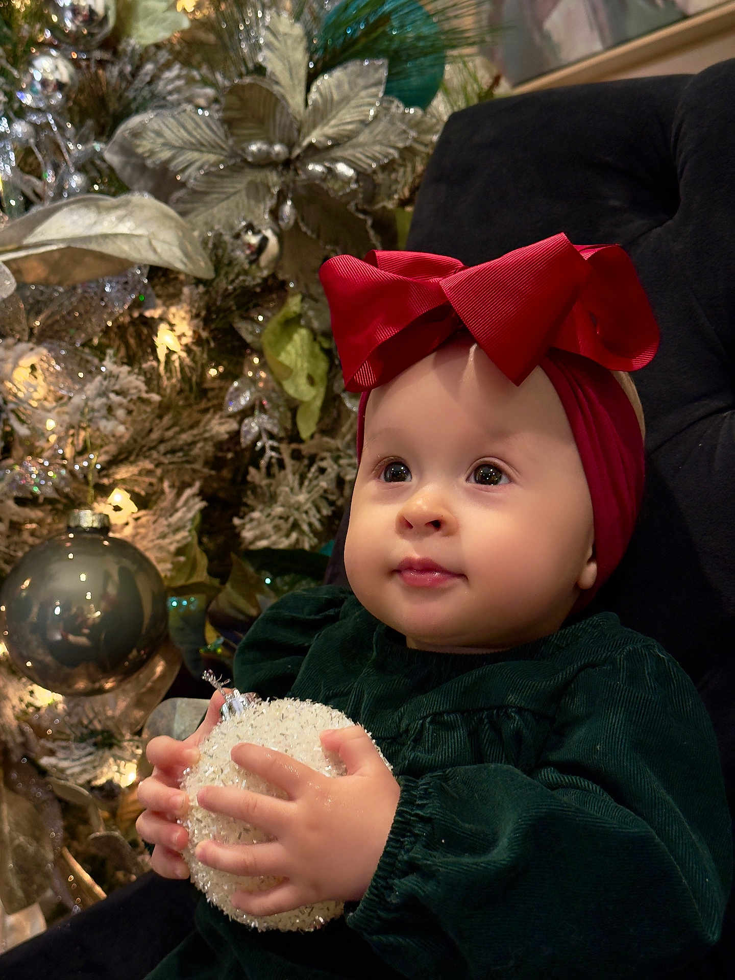 Ainara is registered to the contest to win money with this photo: baby, child, red_bow, christmas_ornament, holiday_decor, christmas_tree, lights, green_dress, plush_chair, festive, indoors, cute, portrait, hands, face, holiday_season, decorations, sparkle, toddler, smile