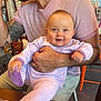 Norah a rejoint le concours — aidez-le/la à gagner de superbes lots ! arm, baby, background_people, beard, cafe, casual_clothing, chair, child, cute, happy, holding, indoor, lavender_onesie, light, man, person, pink_shirt, smile, table, tattoo