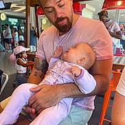 Norah a rejoint le concours — aidez-le/la à gagner de superbes lots ! baby, baby_clothes, background, beard, cafe, casual, chair, child, fun, holding, indoor, interaction, light, man, people, pink_shirt, playful, table, tattoo, tongue_out