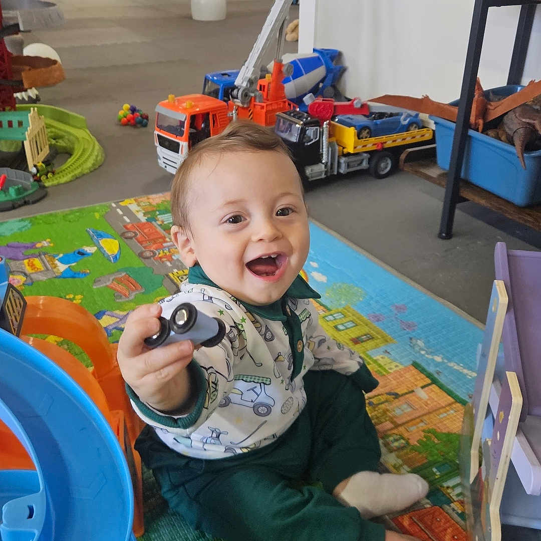 Anthony is registered to the contest to win money with this photo: toddler, child, toy_car, play_mat, slide, toy_truck, indoor, smiling, happy, colorful, floor, plastic_toys, playroom, childhood, fun, young_child, sitting, person, casual_clothing, bright
