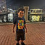 child, boy, birthday_boy, tshirt, shorts, sneakers, brick_pavement, waterfront, night, city_lights, water, smiling, curly_hair, outdoor, urban, reflection, fence, post, happy, portrait