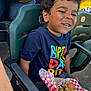 child, smiling, curly_hair, birthday_shirt, burger, food, stadium_seat, green_chair, casual_clothing, happy, person, outdoor, daylight, celebration, fast_food, seat_number, people, hand, jeans, leisure
