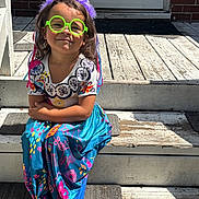 Sarai is registered to the contest to win money with this photo: child, girl, tiara, green_glasses, colorful_dress, sitting, wooden_steps, sunlight, smile, playful, outdoor, patterned_clothing, purple, happy, young, person, casual, fun, portrait, accessory
