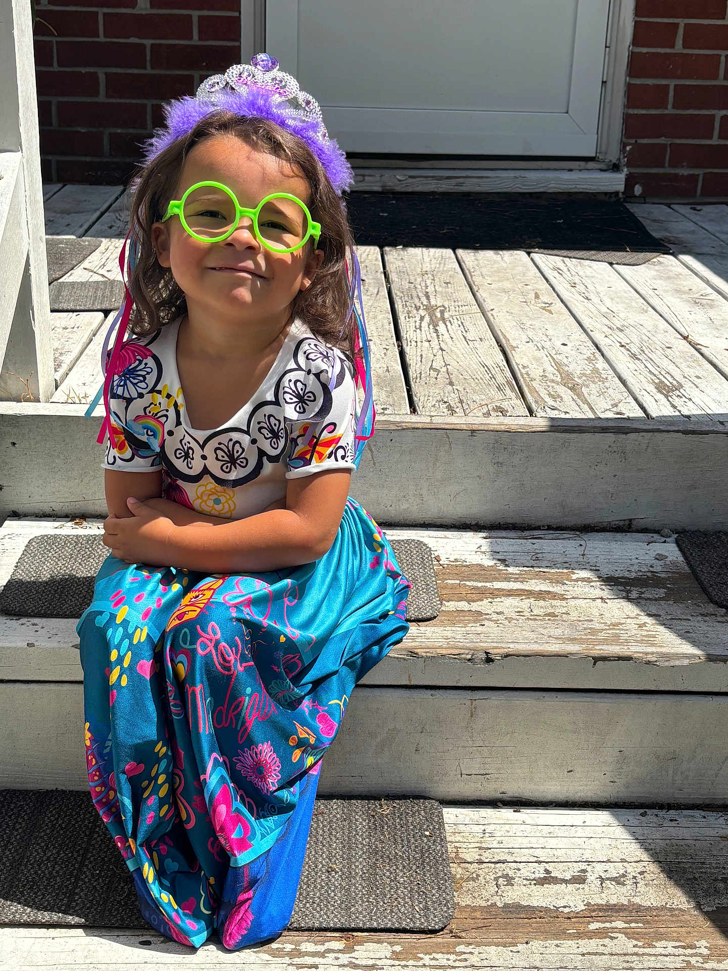 Sarai is registered to the contest to win money with this photo: child, girl, tiara, green_glasses, colorful_dress, sitting, wooden_steps, sunlight, smile, playful, outdoor, patterned_clothing, purple, happy, young, person, casual, fun, portrait, accessory