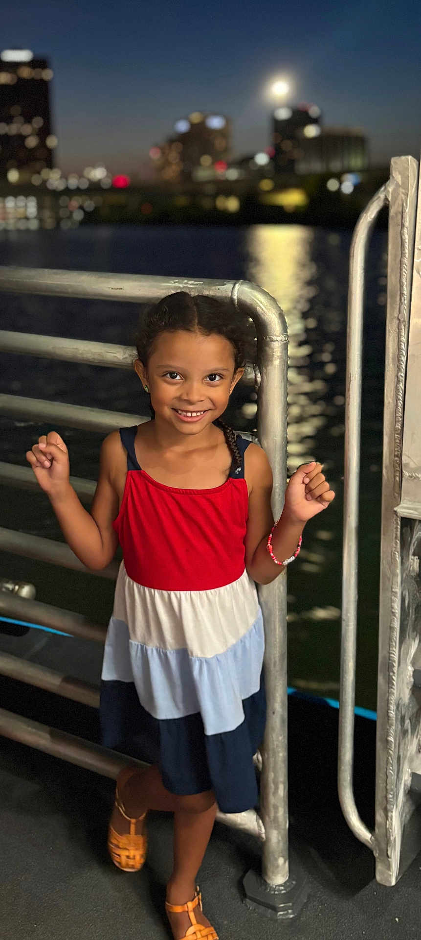 Maleia is registered to the contest to win money with this photo: child, girl, smile, night, city_lights, water, railing, dress, sandals, bracelet, outdoor, urban, portrait, happy, person, fashion, evening, metal, cute, standing