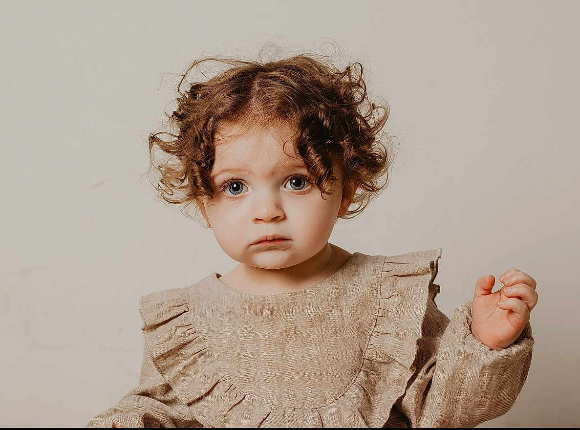 Charlotte joined the competition — help win amazing prizes! toddler, child, curly_hair, blue_eyes, portrait, beige_clothing, ruffles, neutral_background, innocent, serious_expression, soft_lighting, face, person, young_child, studio_photo, cute, fashion, long_sleeves, indoor, closeup