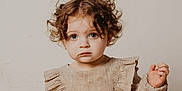 Charlotte joined the competition — help win amazing prizes! toddler, child, curly_hair, blue_eyes, portrait, beige_clothing, ruffles, neutral_background, innocent, serious_expression, soft_lighting, face, person, young_child, studio_photo, cute, fashion, long_sleeves, indoor, closeup