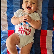 Ezra joined the competition — help win amazing prizes! baby, child, infant, smiling, laughing, onesie, clothing, blanket, quilt, colorful, red, blue, white, happy, cute, playful, person, portrait, indoor, skin