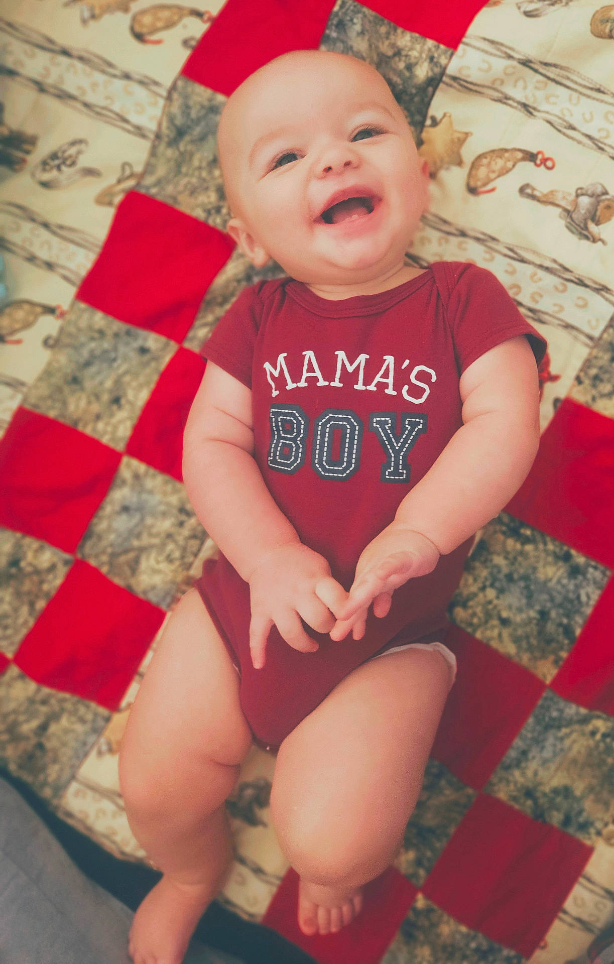 Ezra is registered to the contest to win money with this photo: baby, child, chubby_arms, cute, fabric, face, feet, happy, indoors, infant, legs, lying_down, maroon_clothing, onesie, patchwork_quilt, patterned_background, playful, skin, smiling, text_on_clothing