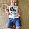 Ezra is registered to the contest to win money with this photo: baby, infant, smiling, tank_top, blue_shorts, bed, blanket, lying_down, happy, cute, child, clothing, person, indoors, casual, small_things_text, skin, feet, hands, face