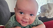 Ezra is registered to the contest to win money with this photo: baby, infant, smiling, face, hands, cushion, red, green_clothing, living_room, sofa, blanket, portrait, cute, head, eyes, skin, indoors, home, person, child