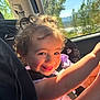 toddler, child, car_seat, smiling, curly_hair, sunlight, window, trees, mountains, car_interior, happy, portrait, seatbelt, playful, daylight, nature, face, person, clothing, travel