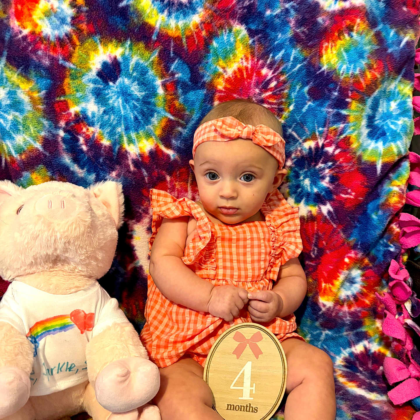 Sophie is registered to the contest to win money with this photo: baby, infant, child, plush_toy, pig, tie_dye_blanket, orange_outfit, headband, wooden_sign, 4_months, colorful, sitting, cute, portrait, indoor, soft_toy, patterned_blanket, smiling, clothing, person