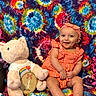 baby, child, infant, smiling, plush_toy, pig, orange_clothing, headband, tie_dye_blanket, milestone, wooden_plaque, sitting, cute, happy, indoors, colorful, portrait, person, fabric, toy