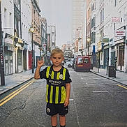Ethan is registered to the contest to win money with this photo: child, boy, soccer_jersey, shorts, sneakers, street, urban, background, pose, gesture, blond_hair, standing, sidewalk, buildings, vehicles, road, lamp_post, casual_clothing, young_person, playful