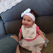 Amaria is registered to the contest to win money with this photo: baby, child, smiling, headband, bib, pink_clothing, sitting, carpet, couch, blanket, foot, person, indoor, happy, toddler, white_bow, sock, denim_jeans, casual, home