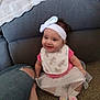 baby, child, smiling, headband, bib, pink_clothing, sitting, carpet, couch, blanket, foot, person, indoor, happy, toddler, white_bow, sock, denim_jeans, casual, home