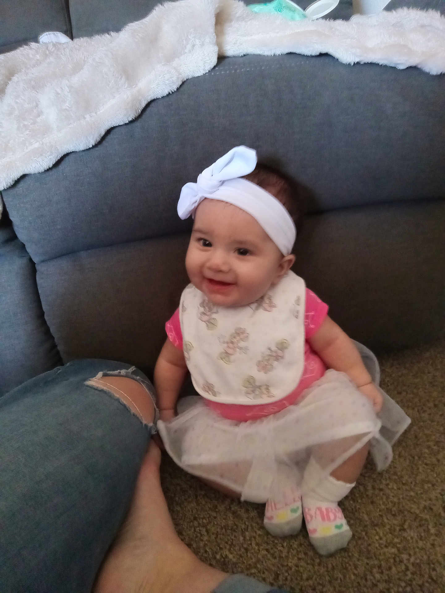 Amaria is registered to the contest to win money with this photo: baby, child, smiling, headband, bib, pink_clothing, sitting, carpet, couch, blanket, foot, person, indoor, happy, toddler, white_bow, sock, denim_jeans, casual, home