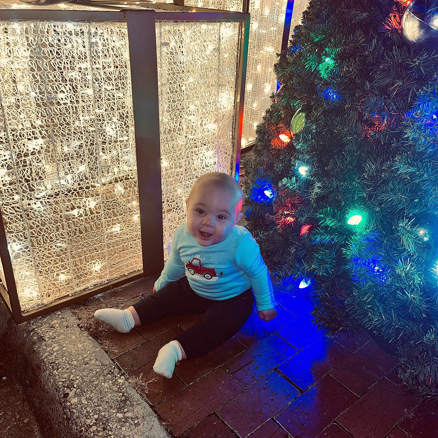 Judson is registered to the contest to win money with this photo: baby, child, christmas_tree, lights, brick_floor, smile, festive_clothing, holiday, decorations, outdoor, happy, sitting, toy_truck_print, sock, night, glowing_lights, pine_tree, person, celebration, winter